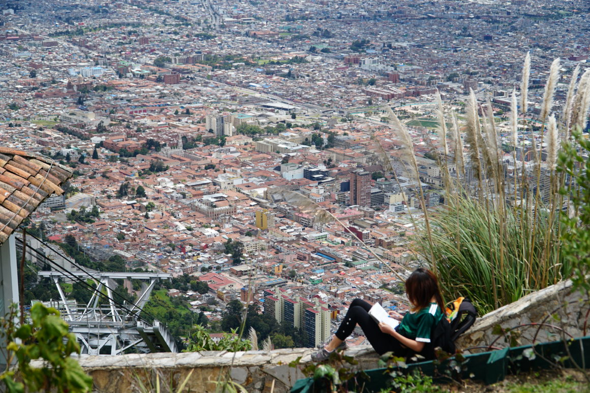 Woman sitting on a ledge, reading a book and the city of Bogota is behind her.